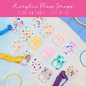 May include: A set of ten rectangular acrylic floss drops featuring various colorful patterns including rainbows, hearts, and floral designs. Each piece has a circular hole and a slide-on label slot. Some drops are wrapped with blue, yellow, or purple embroidery thread.
