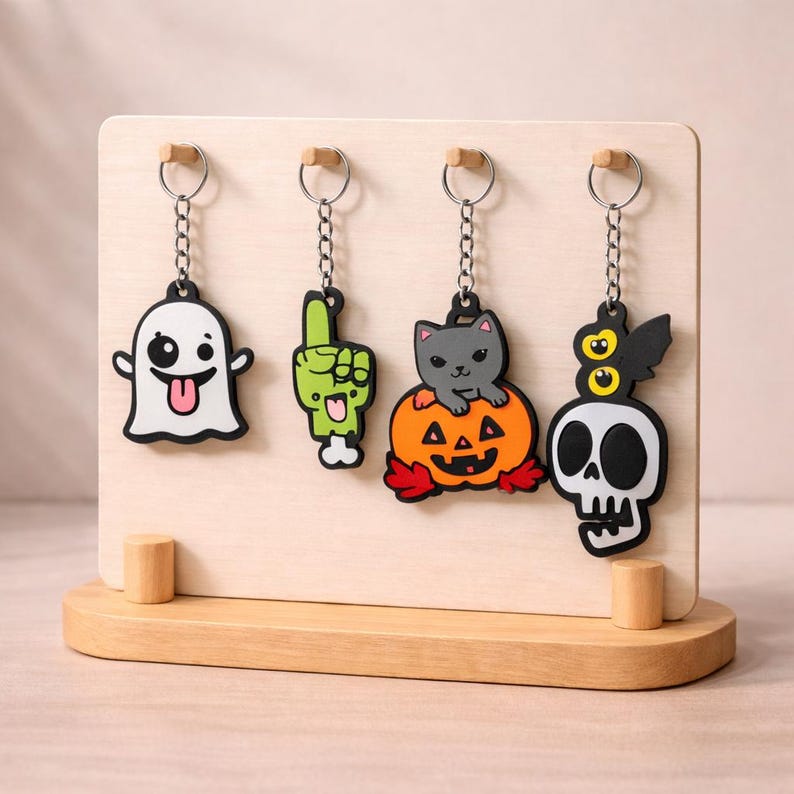 May include: A wooden display stand featuring four Halloween-themed keyrings. The keyrings depict a white ghost, a green hand, a grey cat on a pumpkin, and a skull with bat wings. Each keyring has a silver chain and a wooden top.