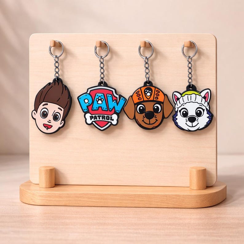 May include: Four colourful Paw Patrol character keyrings displayed on a wooden stand. The keyrings feature Ryder, the Paw Patrol logo, Chase, and Everest. Each keyring has a silver chain and a round metal ring.