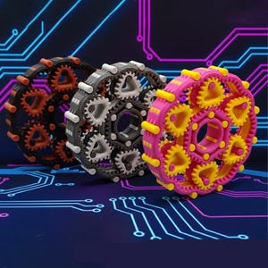 May include: Three colorful gear-shaped objects. One is black with orange gears, another is gray with white gears, and the third is pink with yellow gears. The gears are arranged in a circular pattern, with a central hole.