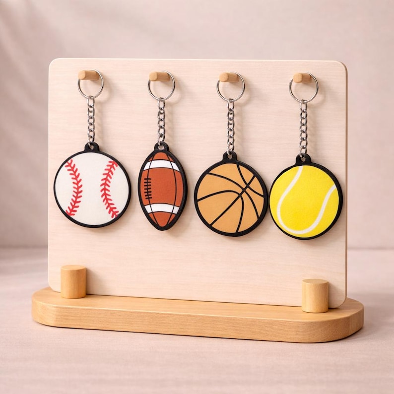 May include: A wooden display stand features four sports-themed keyrings. The keyrings depict a baseball, American football, basketball, and tennis ball. Each keyring has a black border and silver chain. The baseball is white with red stitching.