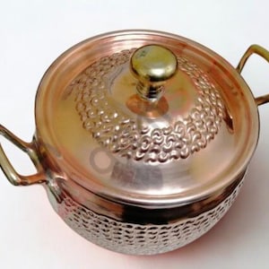May include: A copper cooking pot with a lid. The pot has two handles and a decorative hammered texture on the lower portion. The lid features a textured design and a gold-coloured knob. The pot is round and appears to be made of copper.