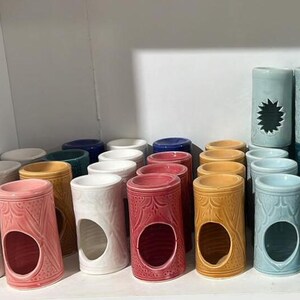 Może przedstawiać: Kolekcja ceramicznych świeczników w różnych kolorach, w tym białym, różowym, czerwonym, żółtym, niebieskim i morskim. Każdy świecznik ma cylindryczny kształt z dużym, owalnym otworem i dekoracyjnymi wzorami.