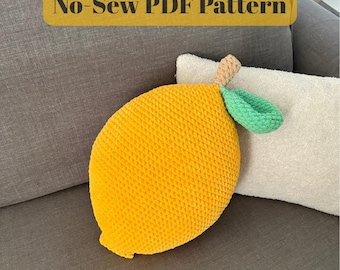 Puede incluir: Un cojín decorativo amarillo con forma de limón con una superficie texturizada, una hoja verde y un tallo marrón. El cojín está sobre un sofá gris con el texto "No-Sew PDF Pattern" y "Lemon Decor Pillow".