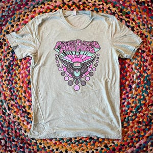 May include: A light green t-shirt with a graphic design featuring the text "Pigeons Playing Ping Pong" in a pink and blue font. The design includes a stylized bird with wings and geometric shapes.