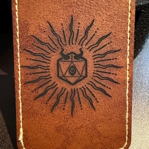 May include: A rectangular brown leather card holder with a white stitched border. The front displays a black sunburst design with a central geometric shape, possibly for a game or fantasy theme.