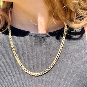 May include: A gold-colored chain necklace with a textured, woven design. The necklace is worn against a black ribbed tank top. The chain is a medium thickness and appears to be made of metal.