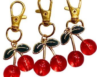 May include: Three cherry keyrings. Each keyring has a pair of translucent red cherry beads, green enamel leaves, and a gold-coloured metal clasp. The keyrings are designed to be attached to keys, bags, or other items.