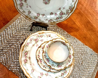 May include: A porcelain dinnerware set featuring a white base with pink floral garlands and gold scalloped edges. The set includes a large dinner plate, a smaller salad plate, a teacup, and a matching plate displayed on a stand.