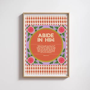 May include: A framed art print with the words "ABIDE IN HIM" in gold lettering. The artwork features a geometric pattern in orange and cream, with a central design of a circle and text, surrounded by pink fruit and green leaves.