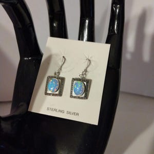 May include: Sterling silver earrings with an oval, opalescent stone set in a square frame. The frame has a decorative border. The earrings are displayed on a white card with the words "STERLING SILVER".