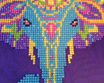 May include: A close-up of a purple fabric with a beaded elephant design. The elephant is made of small, colorful beads in shades of blue, yellow, pink, and purple. The design is centered on the fabric.