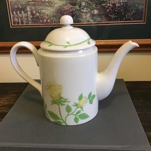 May include: White ceramic teapot with a lid, handle, and spout. The teapot is decorated with yellow rose floral designs and green leaves. A green vine design accents the lid. The teapot is sitting on a gray surface.