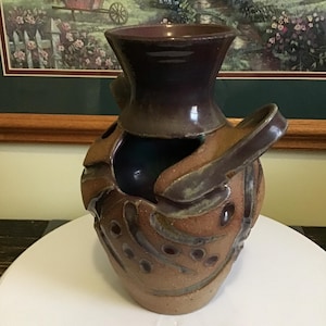 May include: A ceramic vase with a wide, flared neck and two side handles. The body features a textured, light brown clay finish with dark brown, glossy glaze accents and circular indentations. The interior of the vase is a deep, dark purple.