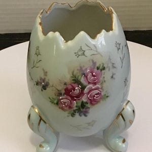 May include: A light blue porcelain egg-shaped vase with a scalloped rim and three curved feet. The vase is decorated with pink roses and gold accents. Delicate floral patterns are painted on the surface.