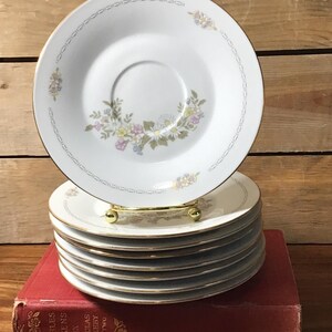 May include: A stack of white ceramic saucers with gold-trimmed edges. Each saucer features a delicate floral pattern of small daisies and pastel-colored blossoms, accented by a thin, repeating geometric border design in a soft grey tone.