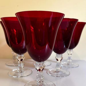 May include: A set of five wine glasses featuring deep red, translucent bowls and clear glass stems with a rounded decorative node above a circular base. The glasses are arranged in a cluster, showcasing the vibrant crimson color of the glass.