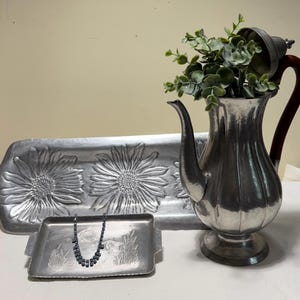 May include: A silver-toned metal coffee pot with a dark wooden handle, a rectangular tray featuring embossed sunflower designs, and a smaller rectangular tray with an etched landscape scene holding a dark beaded necklace with silver accents.