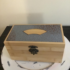 May include: A rectangular wooden box with a light brown finish. The lid features a gray patterned design with a fan-shaped accent. A decorative metal clasp secures the box. The box is on a white plate.