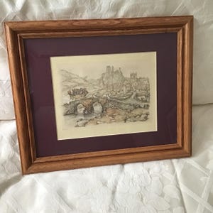 May include: A framed print of a detailed pen and ink drawing depicting a stone bridge, a village, and a castle. The artwork is set against a burgundy mat and encased in a brown wooden frame. The overall scene evokes a sense of history and rural charm.