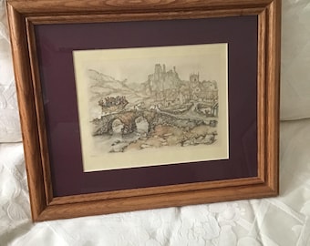Puede incluir: Una impresión enmarcada de un detallado dibujo a pluma y tinta que representa un puente de piedra, un pueblo y un castillo. La obra de arte está enmarcada con un passe-partout burdeos y un marco de madera marrón. La escena evoca historia y encanto rural.