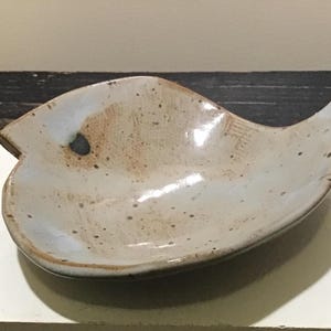 May include: A handmade ceramic dish in a leaf shape. The dish has a speckled, light brown and gray glaze with a dark blue accent. The edges are slightly raised, and the overall design is organic and rustic.