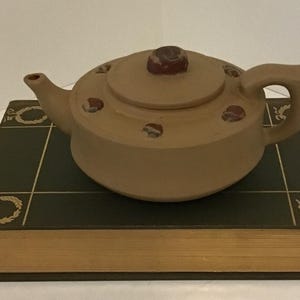 May include: A tan ceramic teapot with a rounded body, spout, and handle, resting on a dark green book. The teapot lid has a dark brown knob and decorative accents. The book has gold-colored page edges and decorative gold trim.