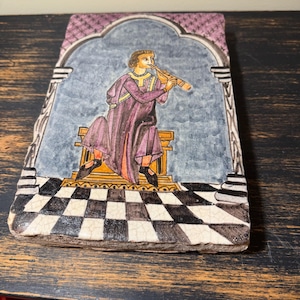 May include: Rectangular ceramic tile with a hand-painted illustration of a figure playing a flute. The figure is dressed in purple robes and sits on a gold-colored platform. The background features a checkered floor and a blue sky.