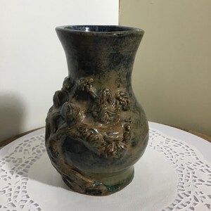 May include: A handcrafted ceramic vase with a textured surface and a dark blue and brown color scheme. The vase features a detailed relief design of a tree and foliage. The vase is a decorative item.