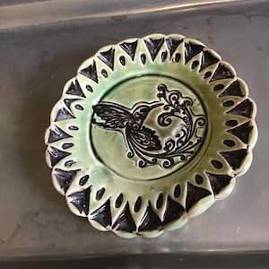 May include: A small, round, ceramic plate with a light green glaze. The plate features a black hummingbird design in the center, surrounded by a decorative border of black triangles. The plate is approximately 5 inches in diameter.