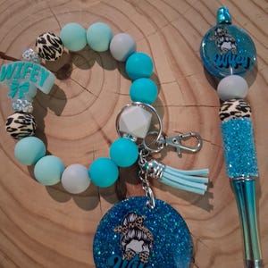 May include: A teal and leopard print beaded keychain bracelet and pen set. The bracelet has a "Wifey" charm and a tassel. The pen has a glittery blue barrel and a matching charm. The keychain has a round charm with a woman's face and the word "Wifey".