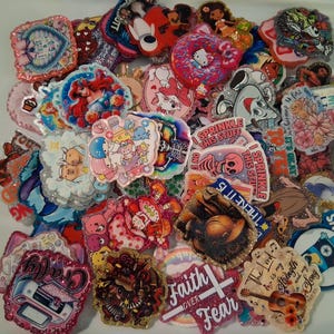 May include: A collection of colorful, glittery resin cabochons with various designs. The cabochons feature cartoon characters, floral patterns, and inspirational quotes. Some have text that reads "Sprinkle This Stuff", "Faith Over Fear", and "Cowboys".