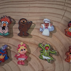 May include: A set of eight colorful enamel pins featuring various Muppet characters including Beaker, Rowlf the Dog, the Swedish Chef, Animal, Gonzo, Miss Piggy, Kermit the Frog, and Fozzie Bear, each with distinct poses and vibrant character-specific colors.