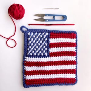 May include: A square crochet potholder featuring a red, white, and blue American flag pattern with a blue border. Included are a ball of red yarn, a silver metal sewing needle, blue metal thread snips, and a red metal crochet hook.