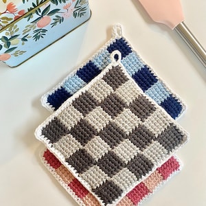 Pode incluir: Três pegadores de panela de crochê feitos à mão com padrões xadrez azuis, marrons e vermelhos. Cada pegador de panela quadrado tem uma borda branca e uma alça para pendurar. Uma espátula rosa e uma lata floral estão no fundo.