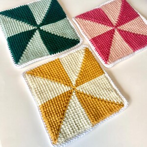 Puede incluir: Tres cuadrados de paños de cocina tejidos a mano. Uno es verde y verde claro, otro rosa y rosa claro, y el tercero amarillo y crema. Cada paño tiene un diseño de molinillo y un borde blanco.