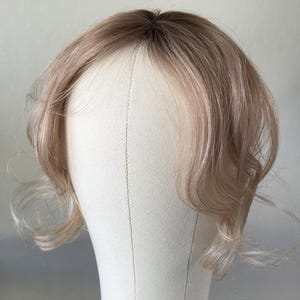May include: A light brown wig with a dark brown root on a mannequin head. The wig has soft, wavy layers and is styled with a side part. The mannequin head is off-white and has a round base.