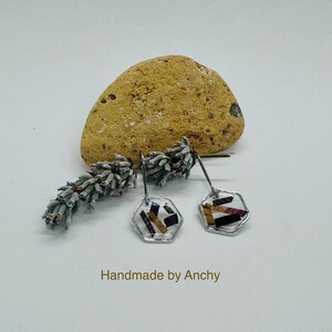 May include: A pair of silver hexagonal earrings with a clear resin design featuring purple and gold pressed flowers. The earrings are handmade by Anchy.