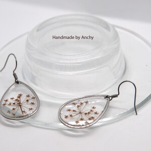 May include: Teardrop-shaped earrings with dried flowers encased in clear resin, set in silver-toned metal. The earrings are displayed on a clear glass plate with a clear glass lid. The text "Handmade by Anchy" is visible.