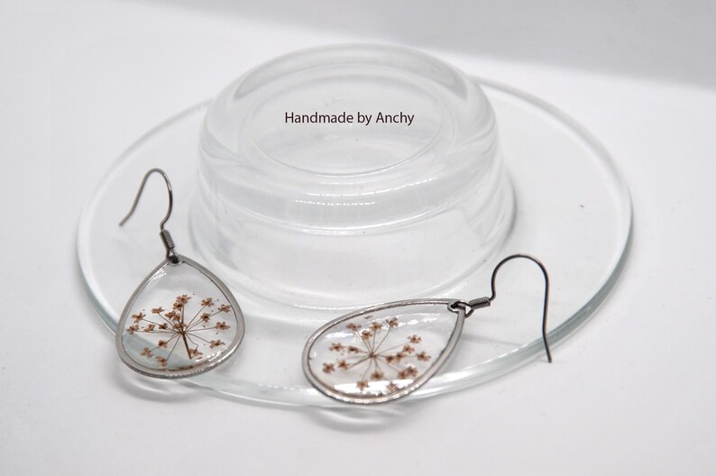 May include: Teardrop-shaped earrings with dried flowers encased in clear resin, set in silver-toned metal. The earrings are displayed on a clear glass plate with a clear glass lid. The text "Handmade by Anchy" is visible.