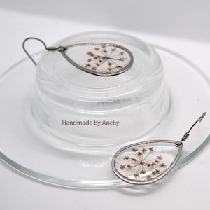 May include: Teardrop-shaped earrings with dried flowers encased in clear resin. The earrings have a silver-colored metal frame and ear wire. The text "Handmade by Anchy" is visible on the glass surface. The earrings are displayed on a clear glass dish.