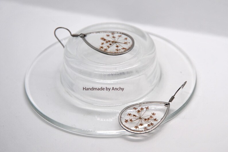 May include: Teardrop-shaped earrings with dried flowers encased in clear resin. The earrings have a silver-colored metal frame and ear wire. The text "Handmade by Anchy" is visible on the glass surface. The earrings are displayed on a clear glass dish.