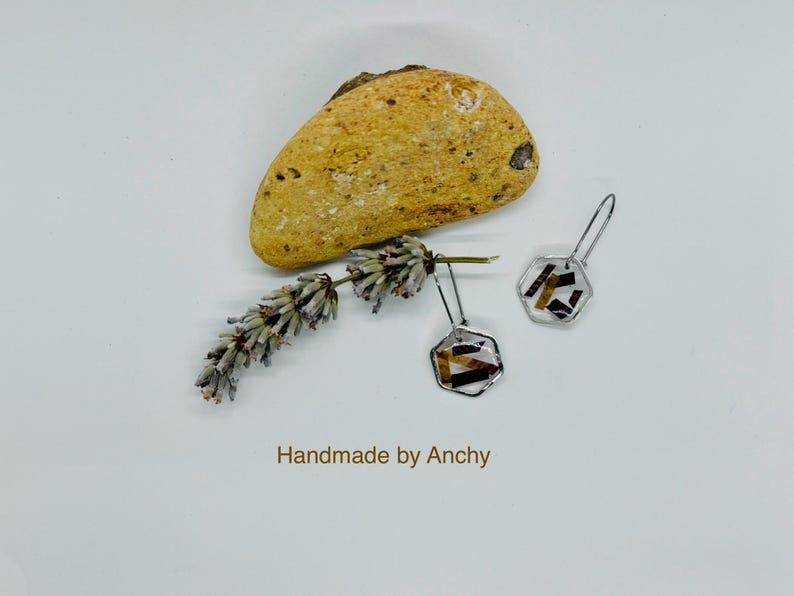 May include: A pair of silver earrings with hexagonal resin accents. The resin is clear and contains small pieces of brown and red material. The earrings are hanging from a white surface with a small rock and a sprig of lavender.