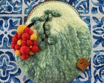 May include: A round felt coin purse in shades of light and dark green, featuring a cluster of red and yellow felted spheres and green leaves. The purse has a metallic frame with two heart-shaped clasps and a small brown leather tag labeled Handmade by Anchy.