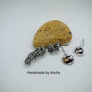 May include: A pair of silver hexagonal earrings with a geometric pattern of brown, black, and white inside. The earrings are on a white surface with a small piece of dried lavender and a yellow rock.