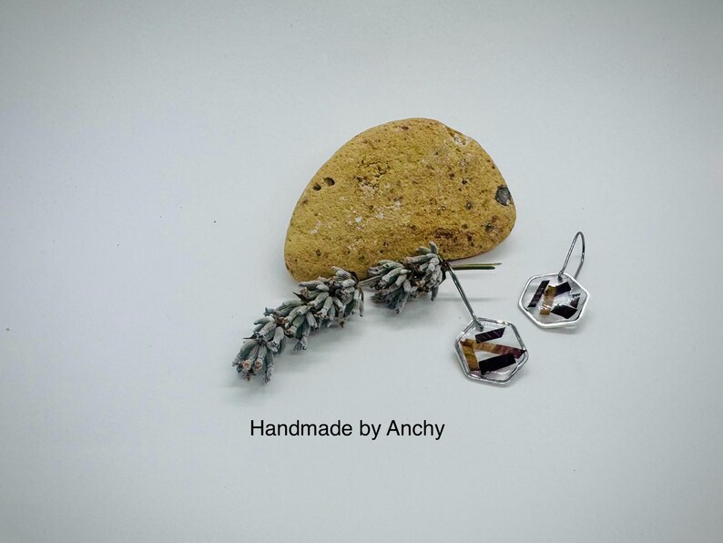 May include: A pair of silver hexagonal earrings with a geometric pattern of brown, black, and white inside. The earrings are on a white surface with a small piece of dried lavender and a yellow rock.