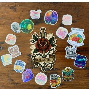 May include: A collection of colorful stickers with various designs and text. Stickers include a brain, a rose with a syringe, and phrases like "Healing Hurts" and "Straight Outta Clinicals." The stickers are arranged on a wooden surface.