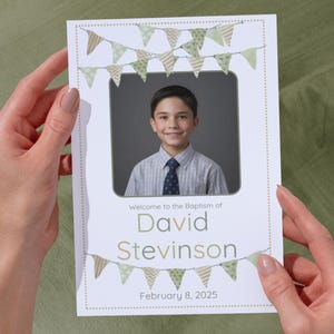 May include: A baptism announcement card held by hands. The card features a photo of a smiling boy, with text that reads "Welcome to the Baptism of David Stevinson" and the date "February 8, 2025". The card is decorated with green and tan pennant banners.