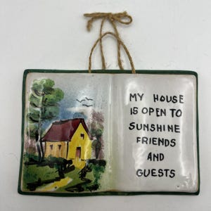 May include: A rectangular ceramic wall plaque shaped like an open book with a dark green border and a jute twine hanger. The left page features a painted yellow house with a red roof, trees, and birds. The right page displays the black text: MY HOUSE IS OPEN TO SUNSHINE FRIENDS AND GUESTS.