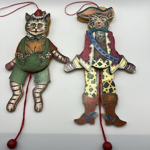 May include: Two articulated wooden cat ornaments with red pull strings. The left cat wears a green outfit and a red hat. The right cat is dressed as a pirate in a red coat, yellow patterned pants, and a hat, featuring a blue sash with heart details.
